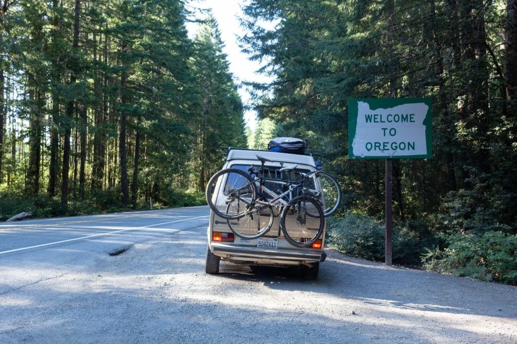 The back of a yellow VW van with 2 bikes near to a "welcome to Oregon" sign