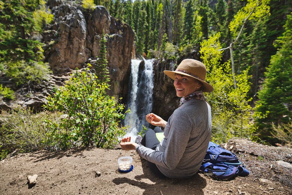 Woman sitting at edge of rock ledge overlooking waterfall eating lunch