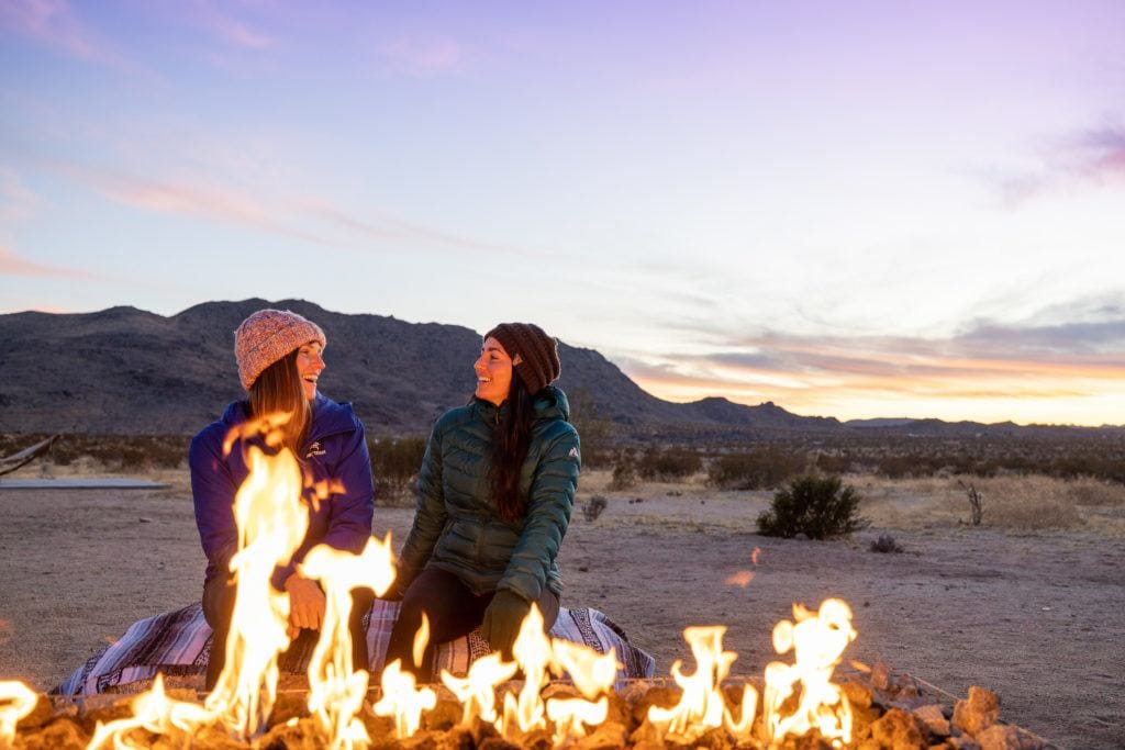 Duas mulheres sorriem uma para a outra sentadas ao lado de uma fogueira no Parque Nacional Joshua Tree