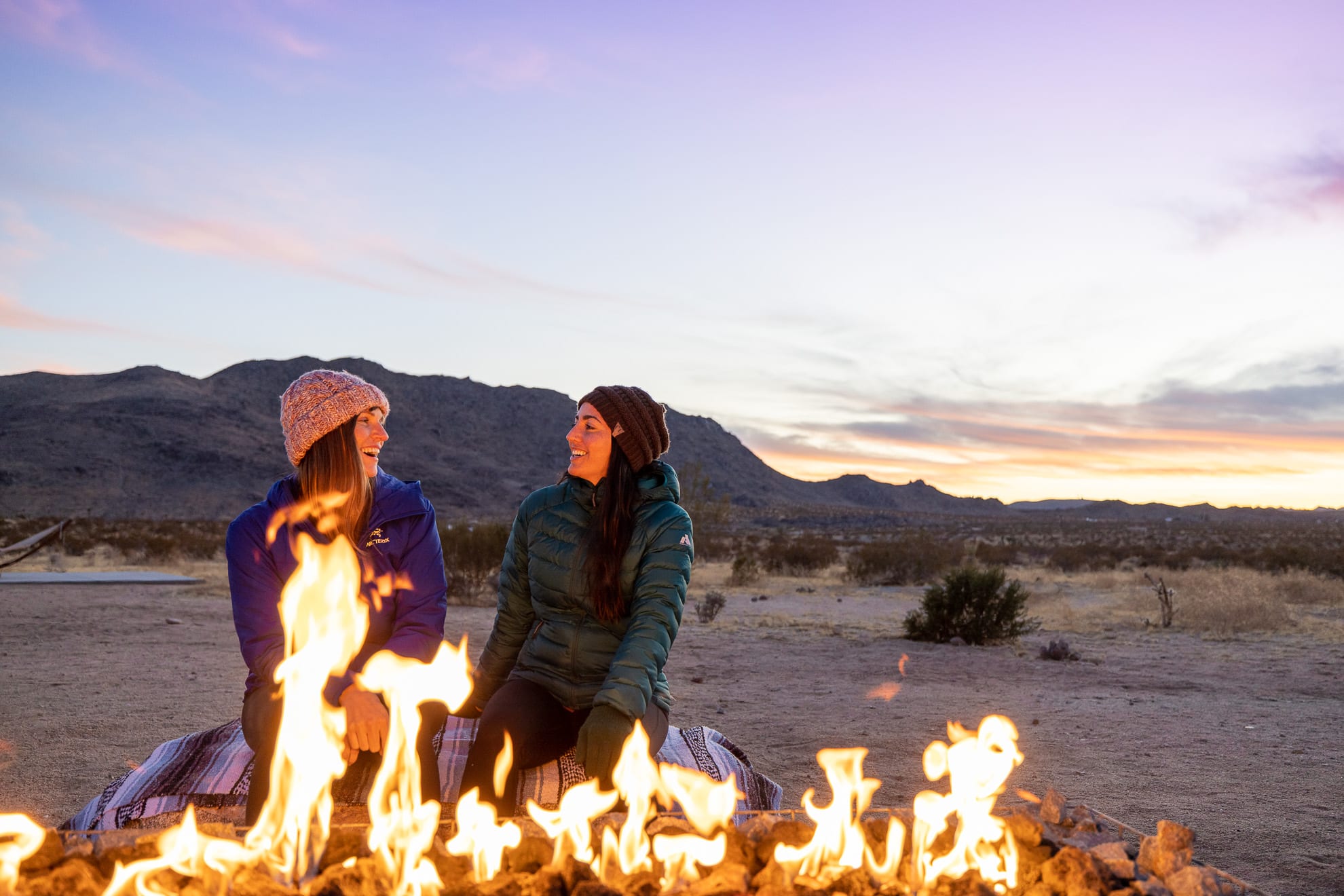 Two women smile at each other sitting next to a campfire in Joshua Tree National Park