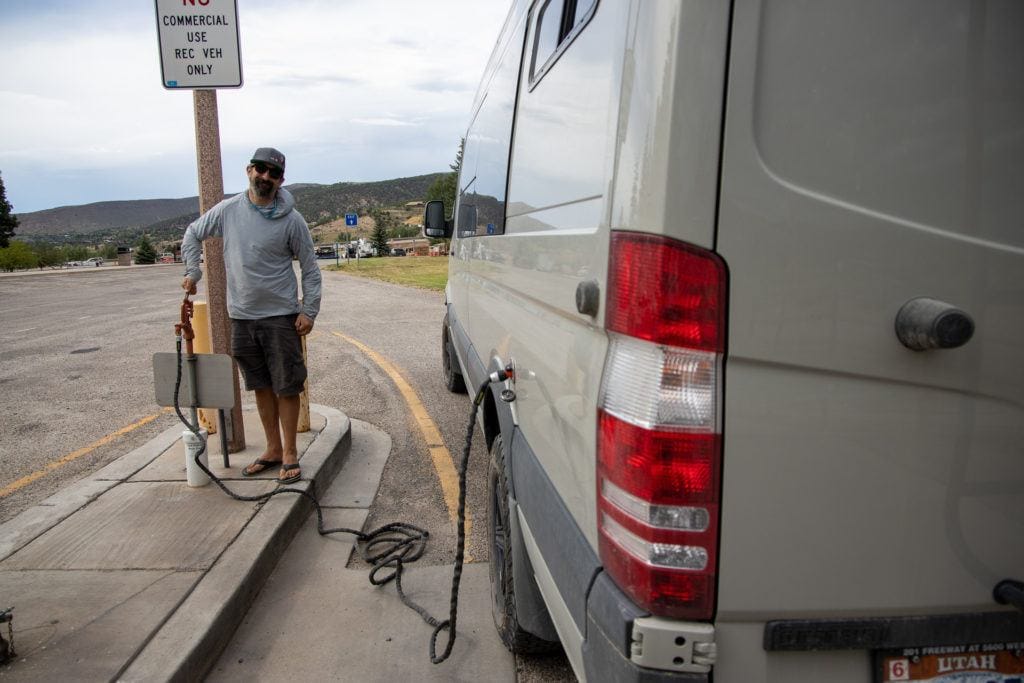 A man stands next to a water pump while refilling a Sprinter water tank at a campground, one of the best places to refill water in van life