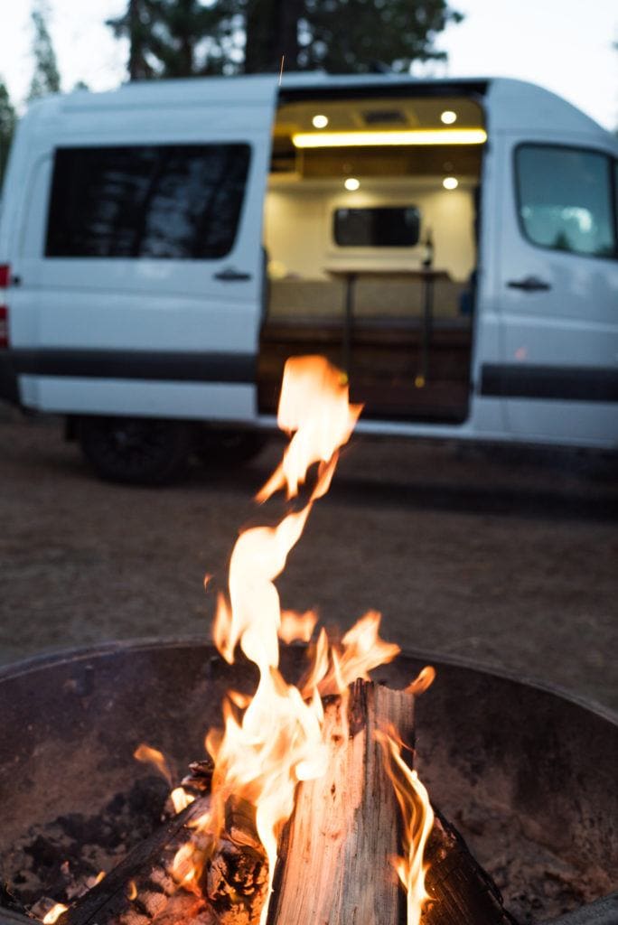 Close up of campfire in metal fire ring. Camper van with open slider door and lights on in background