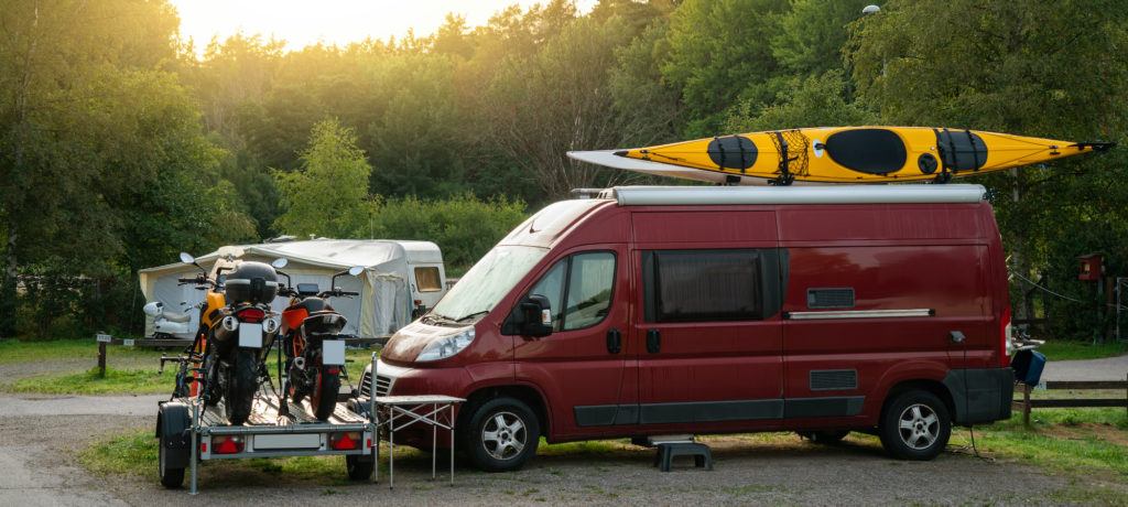 Camper van with kayak on roof and motorbikes on trailer in front of van parked a campsite