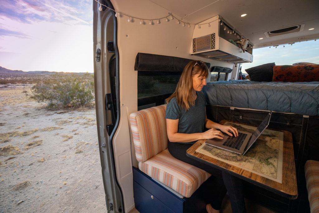 A woman sits in a Sprinter van working on her laptop with the sliding door open
