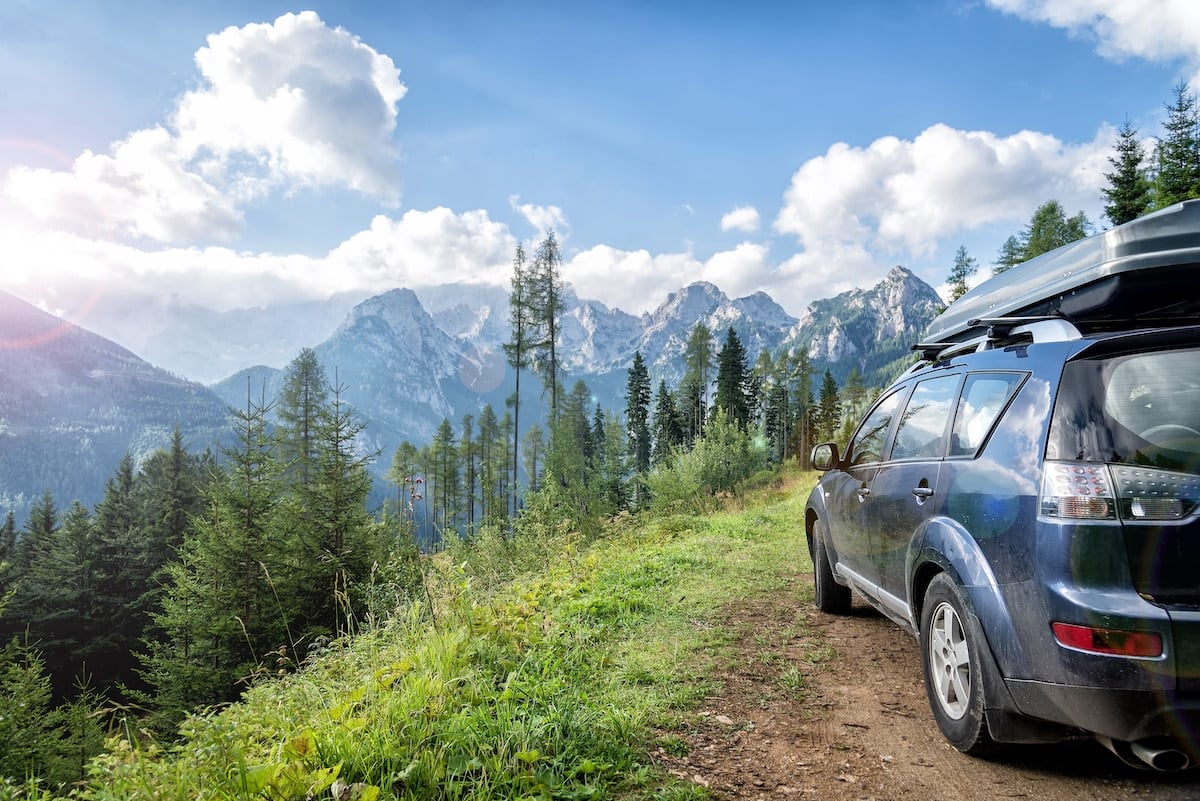 A blue sedan is pulled over on the side of a dirt road on a road trip. There are snowcapped mountains and pine trees in the distance.