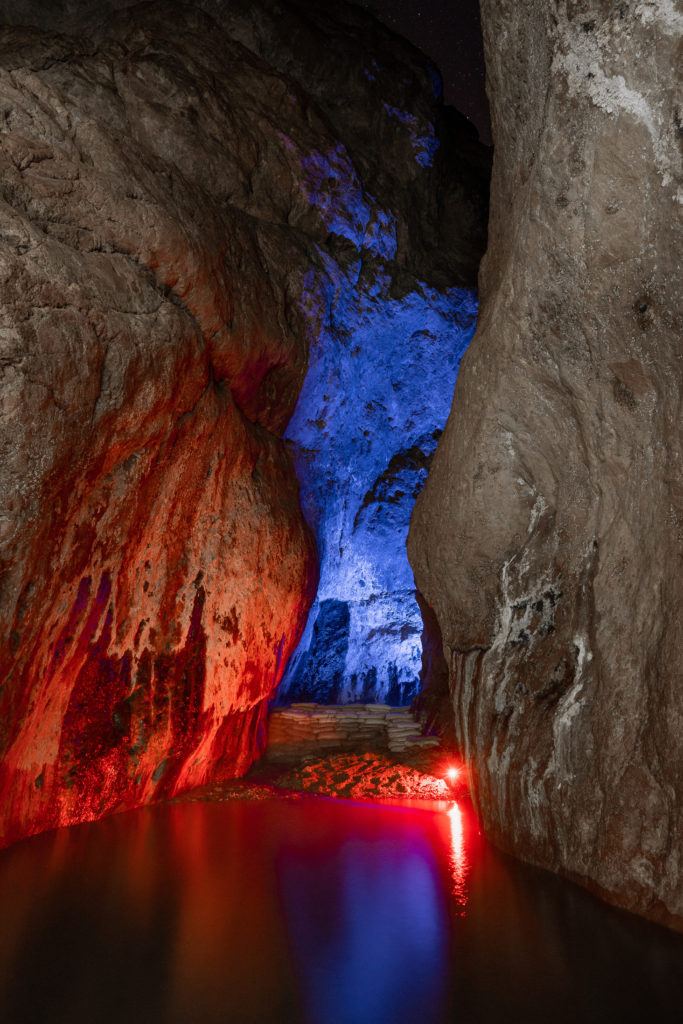 Narrow rock tunnel with pool of water lit with red and blue lights