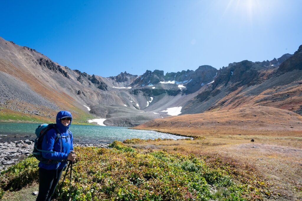 Kristen Bor wearing hiking gear standing next to a blue mountain lake with snow capped mountains in the background