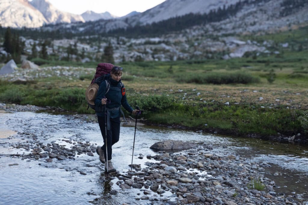 Kristen Bor wearing waterproof hiking boots and backpacking gear walking across a shallow stream