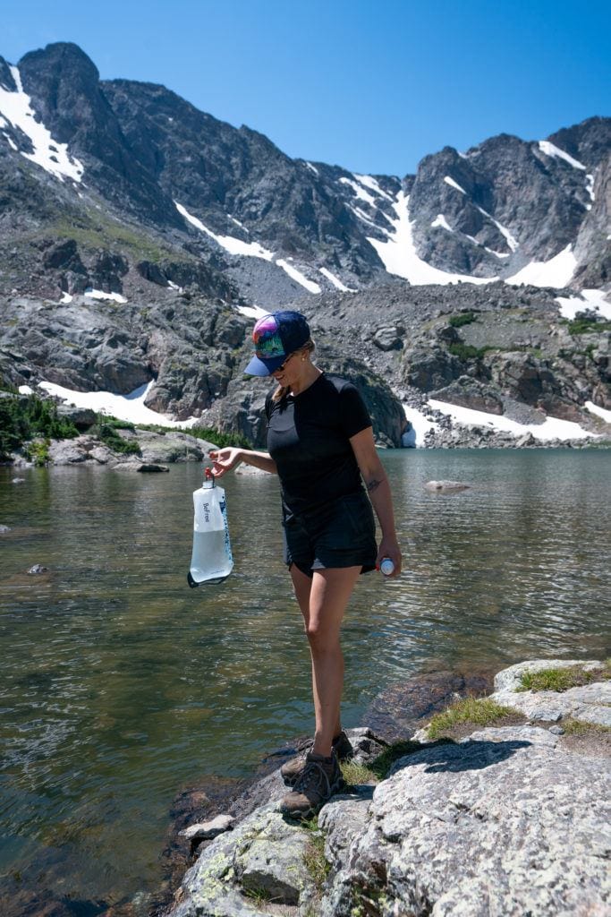 Kristen walking on rock slab a lake water edge carrying water filter filled with water with mountains covered with patches of snow in background