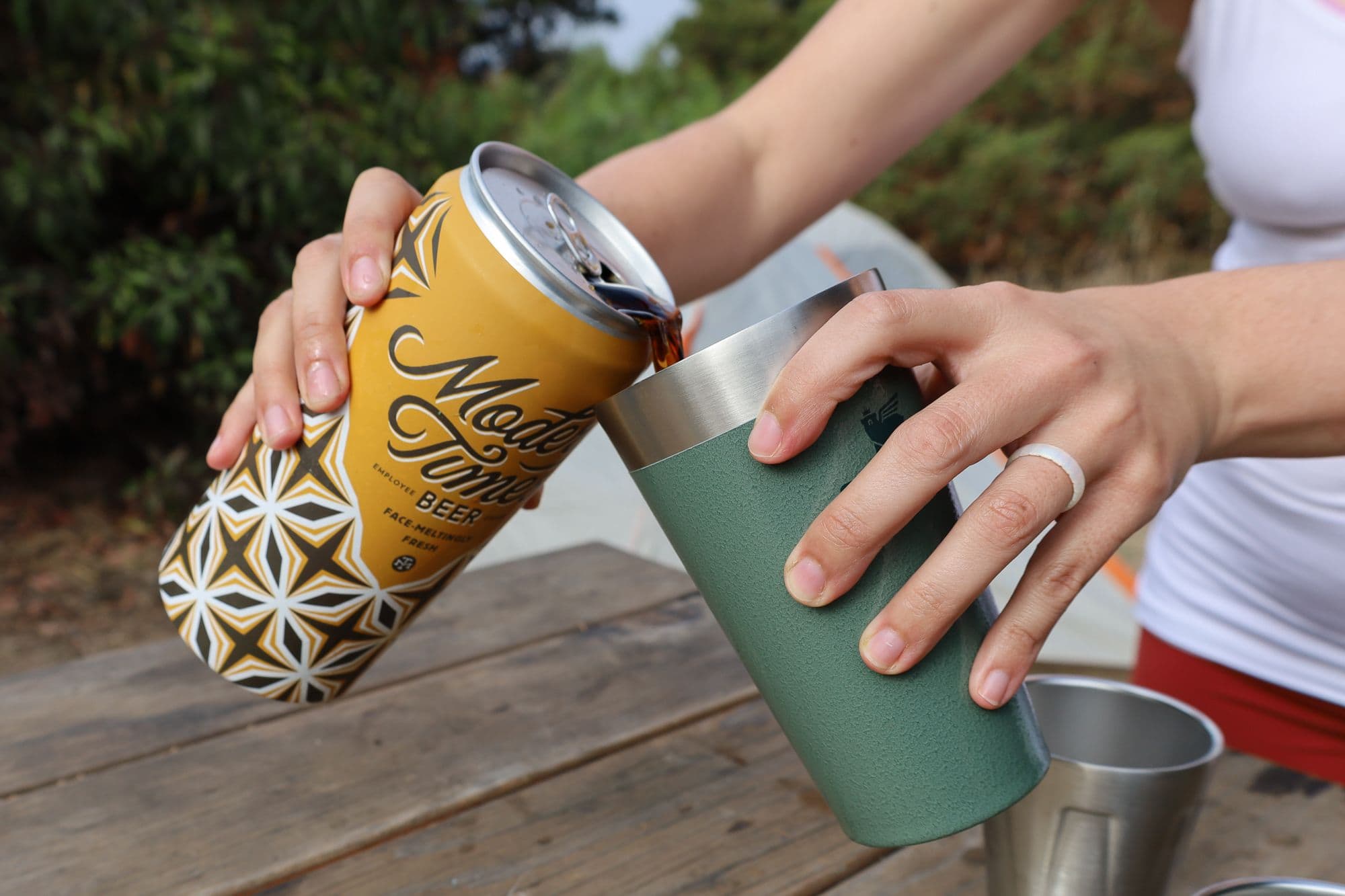 A woman's hands pouring a can of beer into a green pint glass at a wooden table at a campsite