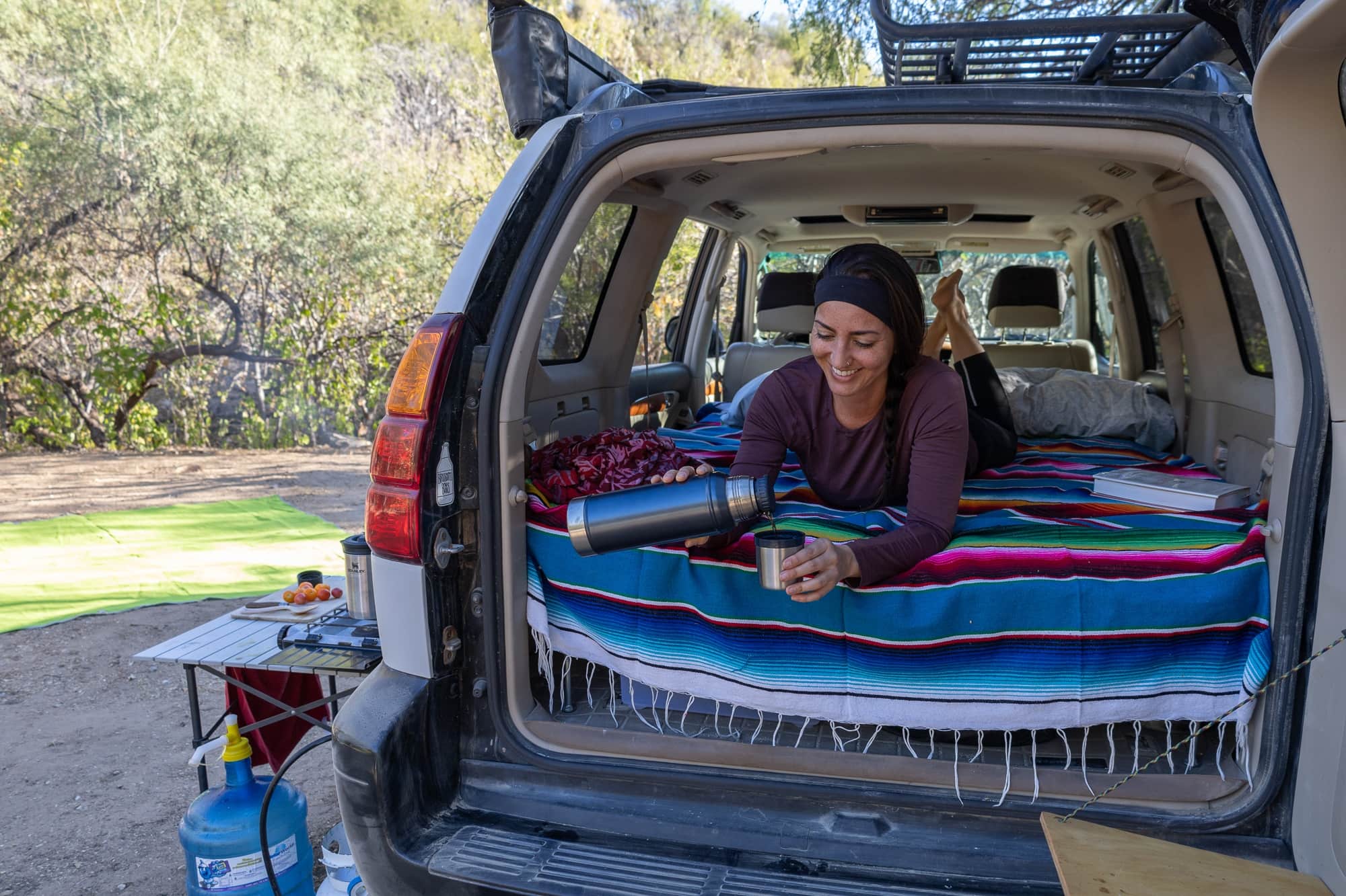 Woman pouring coffee from a thermos into a cup while laying in the back of a car camping vehicle with a blanket