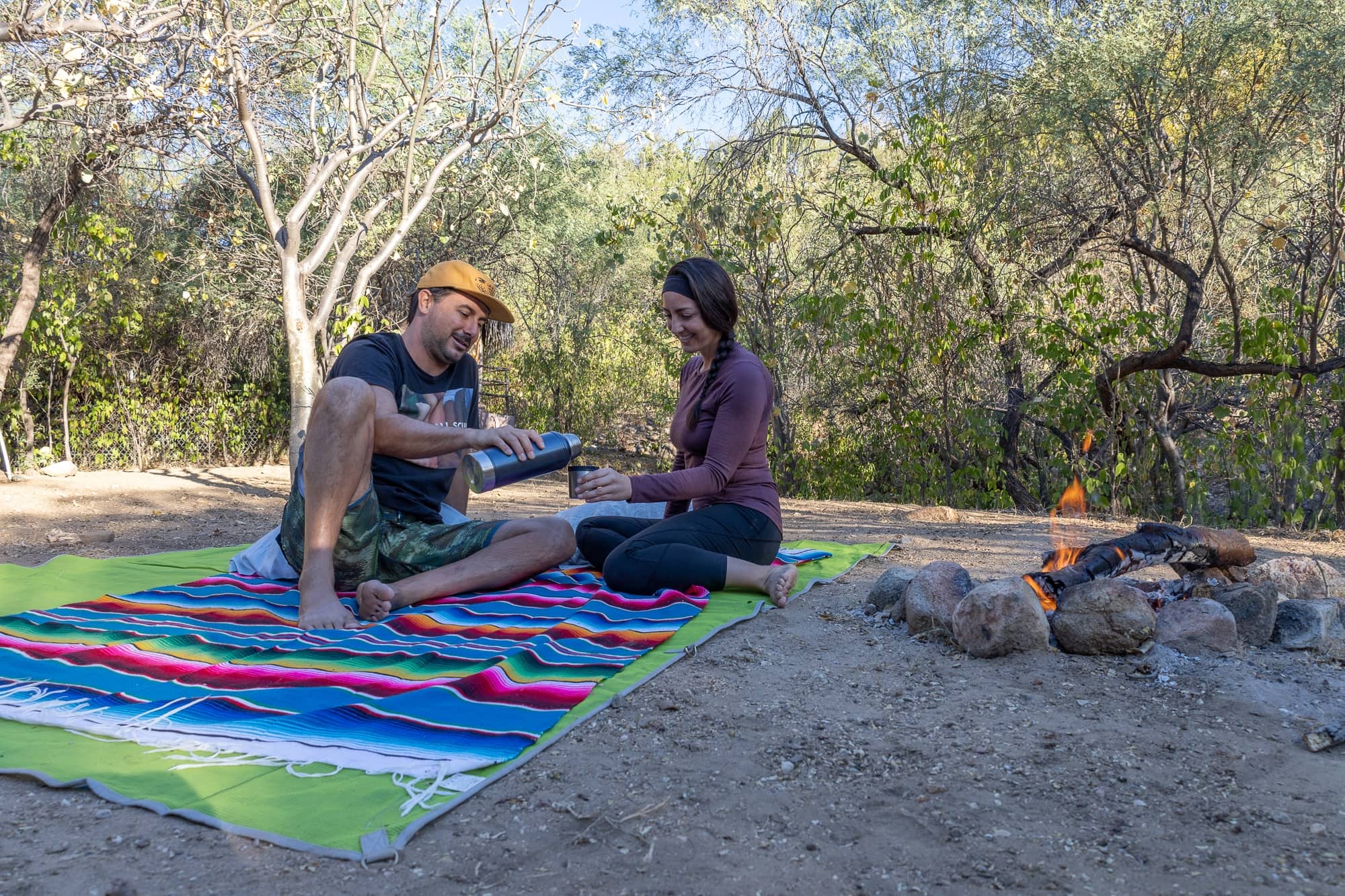 A man pouring a woman a drink form an insulated thermos on a blanket at a campsite with a fire.