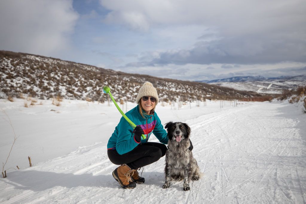 Kristen crouched down next to dog on snowy trail in Utah wearing winter hiking gear