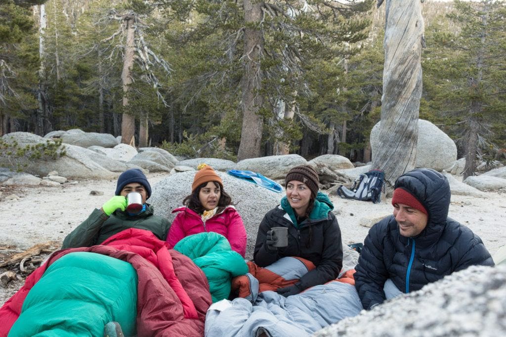 Group of four campers sitting next to each other on ground bundled in warm clothing and sleeping bags