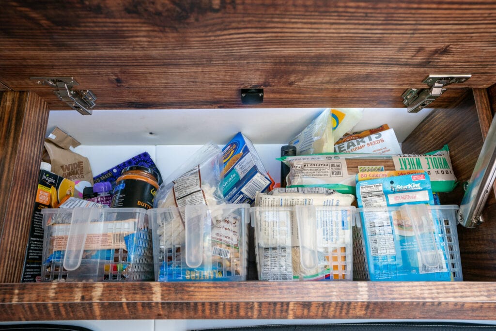 The inside of a cabinet in a Sprinter Van with plastic bins full of shelf stable food