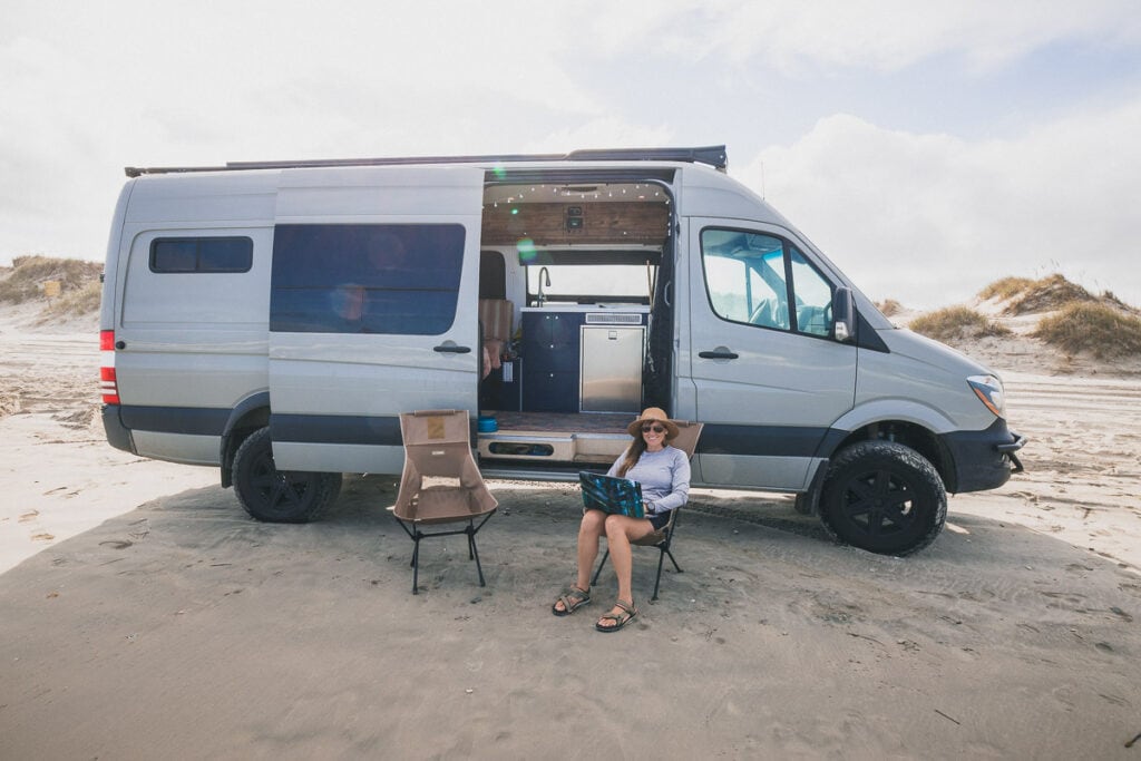 Kristen Bor sitting in a Helinox Camp Chair on the beach next to her Sprinter van with a computer in her lap