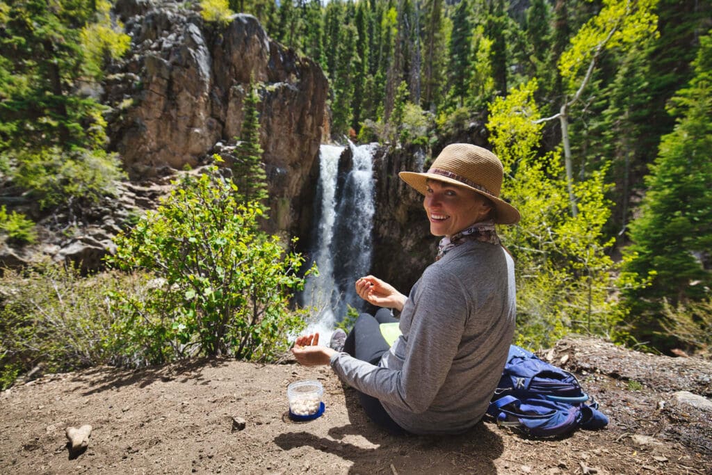 Woman eating nuts by a waterfall // Learn how to train for hiking with easy tips and tricks so you can get in shape and be prepared for your next hiking adventure.