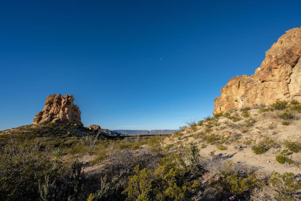 A scenic desert landscape featuring rocky terrain with scrubby vegetation. Two large rock formations are prominent, one on the left and the other on the right, under a clear blue sky with a small distant moon visible.