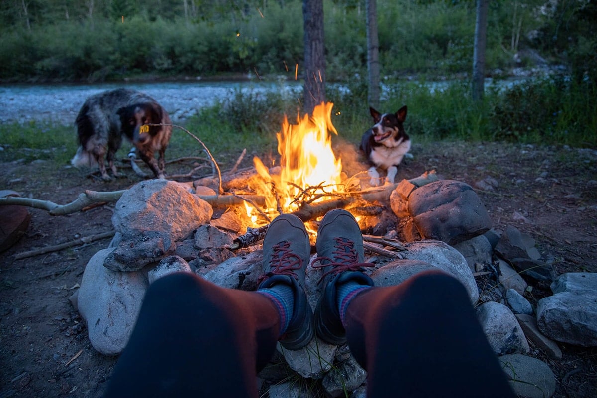 Woman's feet in hiking boots propped on a rock around a campfire with two dogs behind it