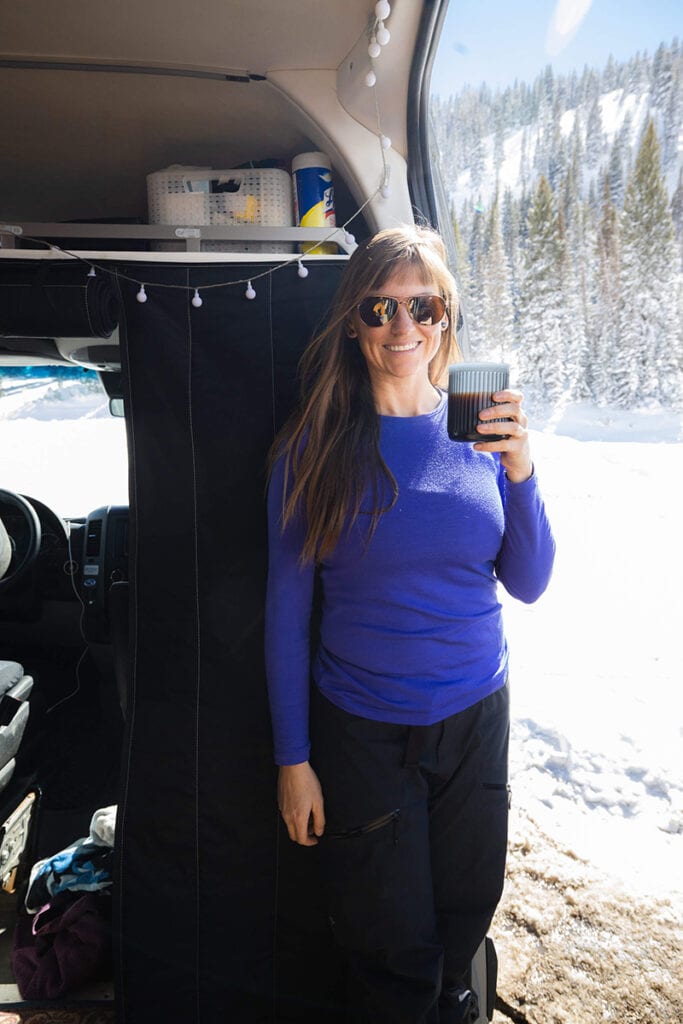 A woman smiles standing in the doorway of her Sprinter van drinking a cup of coffee made by the Aeropress Go