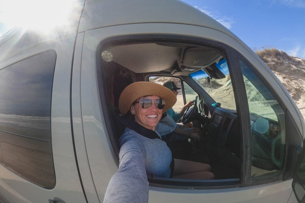 A woman takes a selfie out the window from the passenger seat on a road trip