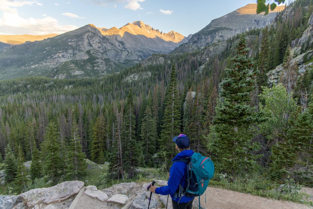 a woman wearing a jacket and daypack looks out toward a mountain with lots of trees in the distance