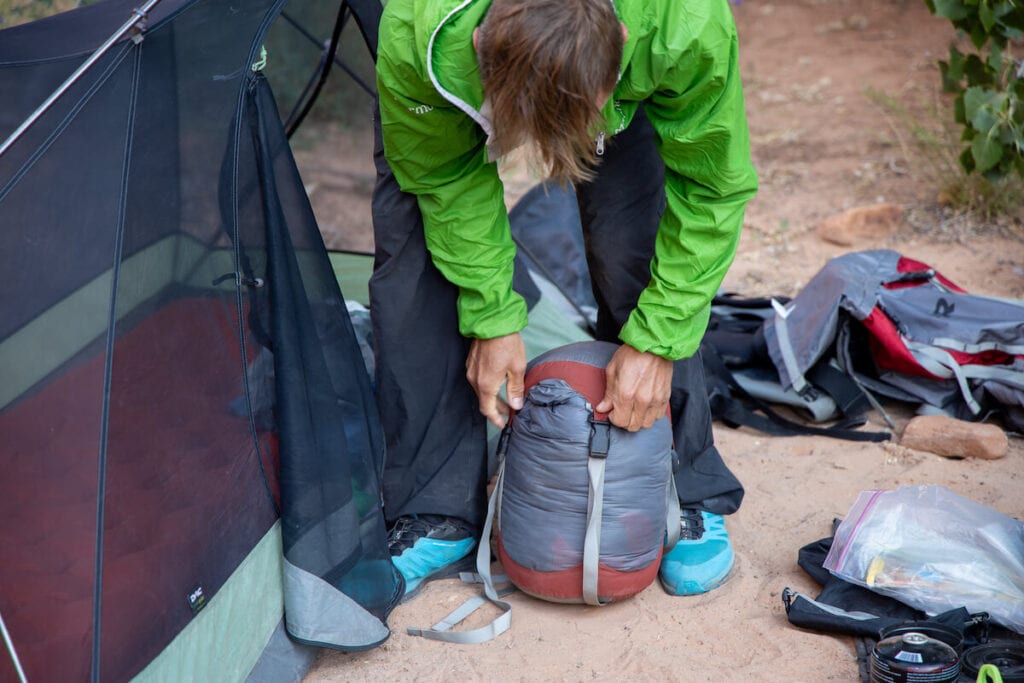 a man stuffing a sleeping bag into a compression sack next to his tent on a backpacking trip