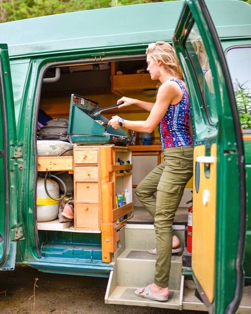 Woman cooking on a propane stove in a budget DIY green Ford Econoline camper van 