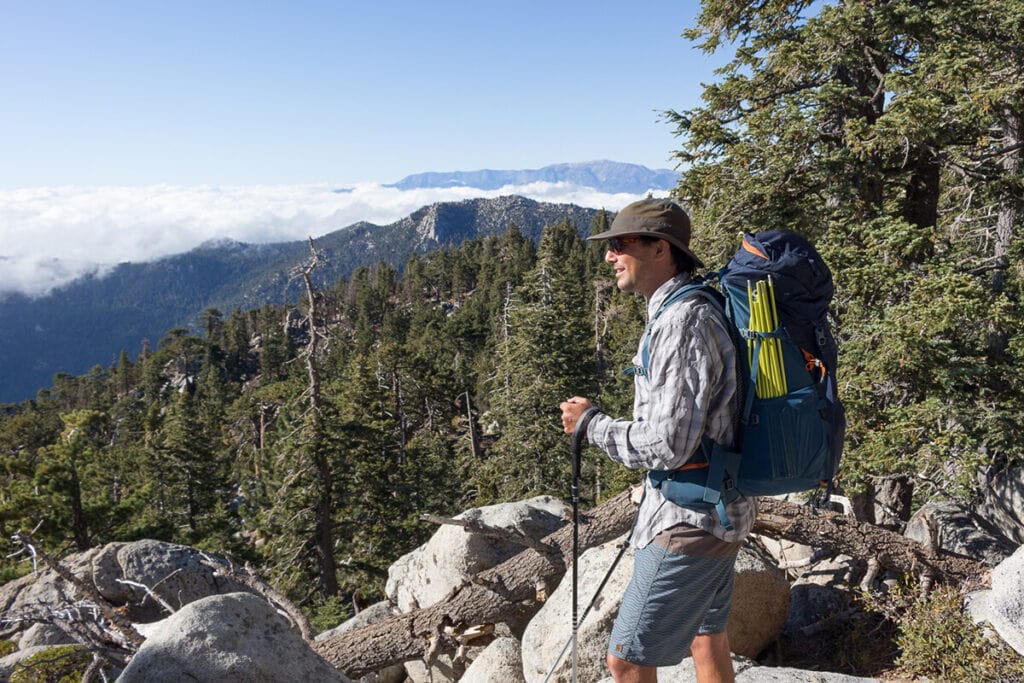 A man smiles standing to the side of the camera. He's wearing a backpacking pack and hiking San Jacinto Peak