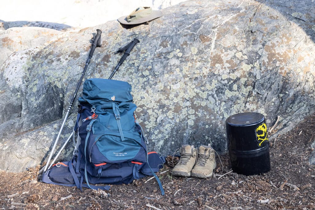 Backpacking gear including a backpacking pack, trekking poles, hat, hiking boots, and a bear canister