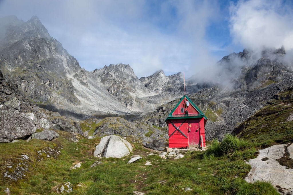 Small, red Alaska Backcountry Hut with rocky jagged mountains as backdrop