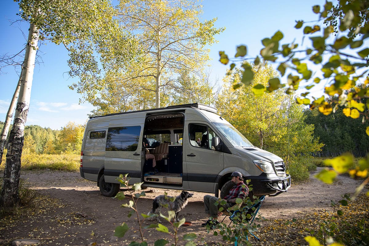 A man sitting in a camp chair with a dog in front of a converted Sprinter van in Colorado surrounded by golden fall aspens