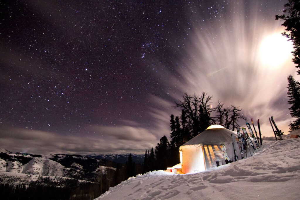 Backcountry winter hut lit up with lights overlooking snow-covered ridges and mountains