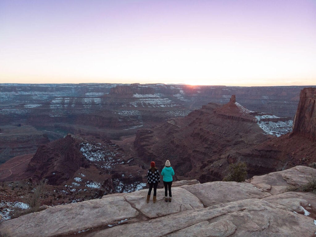 Two women standing facing sunset at Dead Horse Point in Moab. Red rocks have a scattering of snow