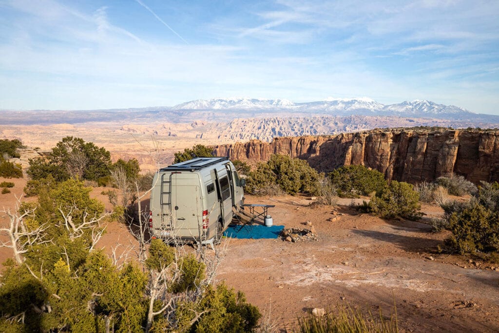 A Sprinter van at a dispersed campsite in Moab, Utah with a camp chair, rug, and table set up outside