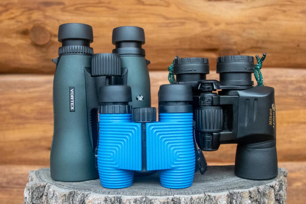 Three pairs of hiking binoculars on a stump with a wood cabin background