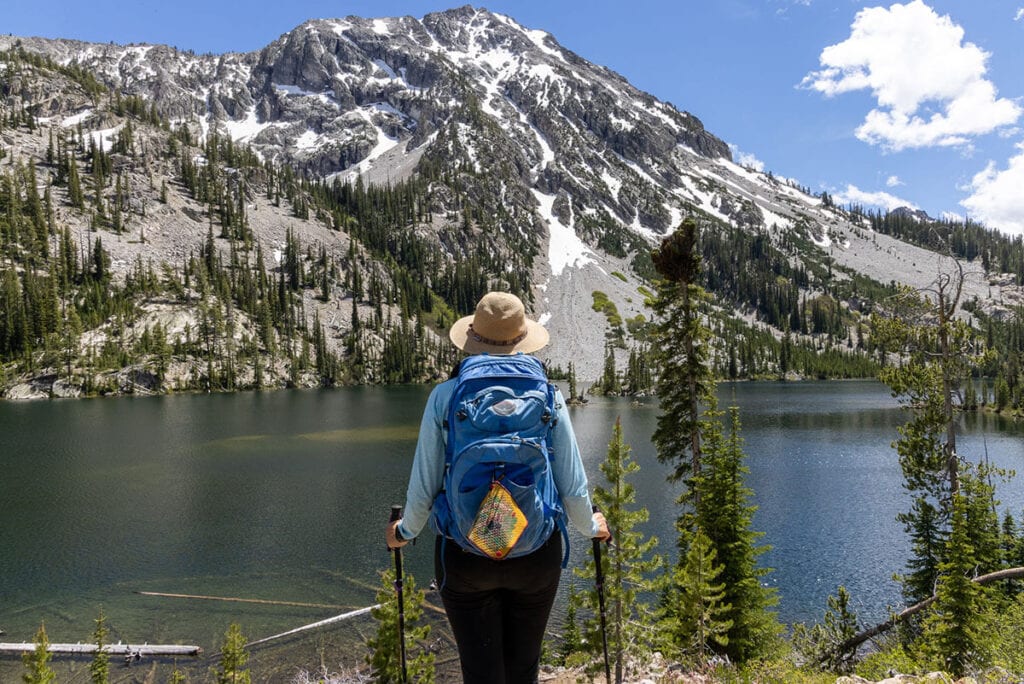 Hiker facing away from camera looking out onto alpine lake with snow-dusted peak in background