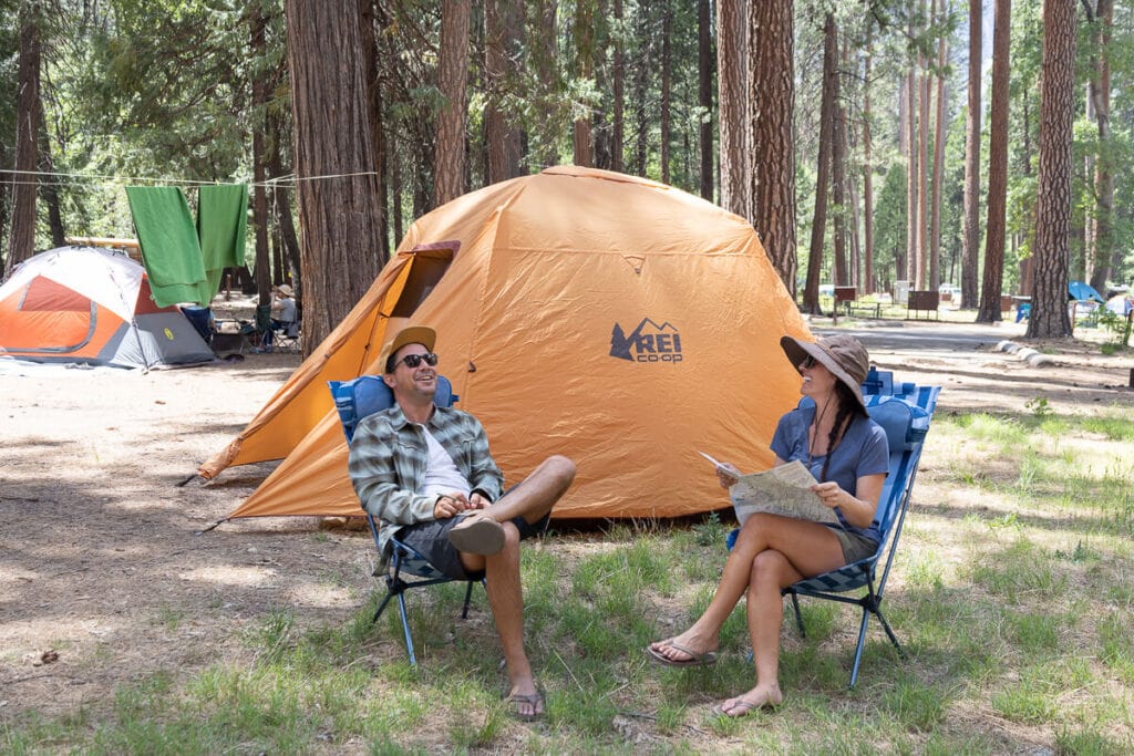 Two people sit in the Helinox Sunset Camp Chairs in front of an REI tent