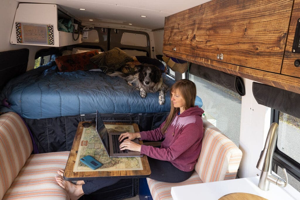 Kristen working on her laptop inside her converted Sprinter camper van with her dog laying on the bed next to her