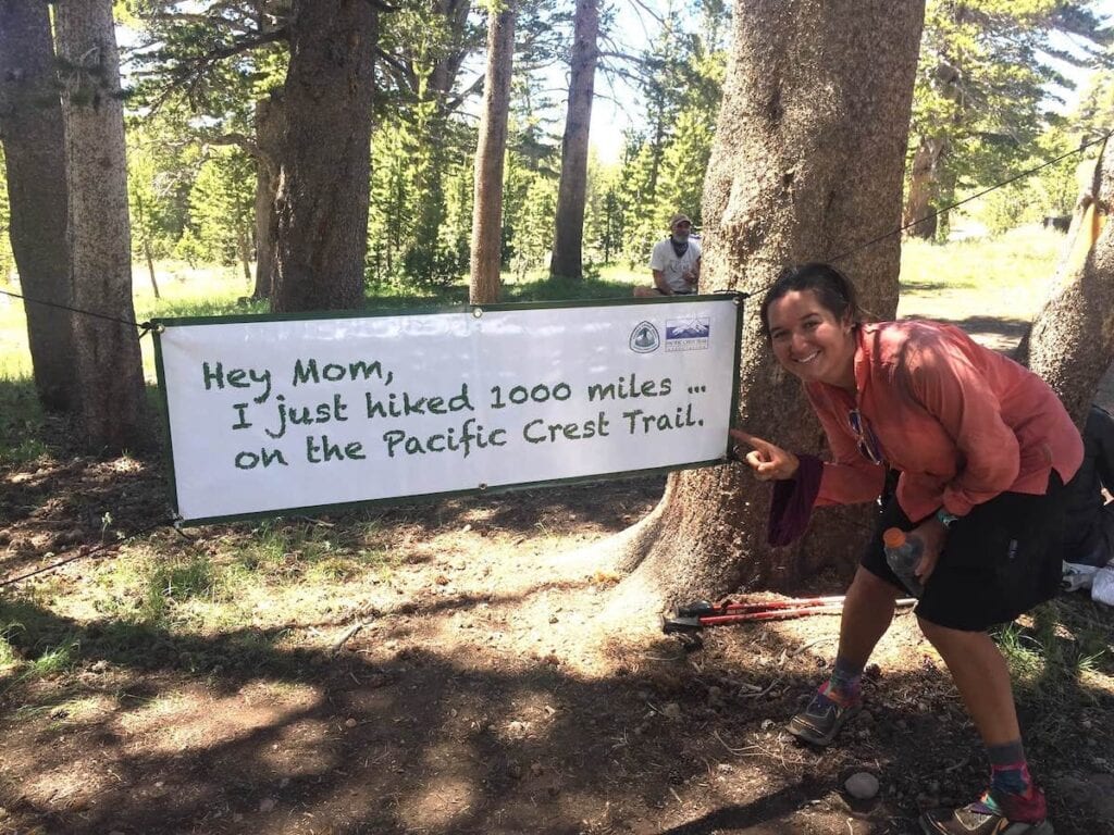 Woman standing next to sign that says "Hey mom, I just hiked 1000 miles on the the Pacific Crest Trail"