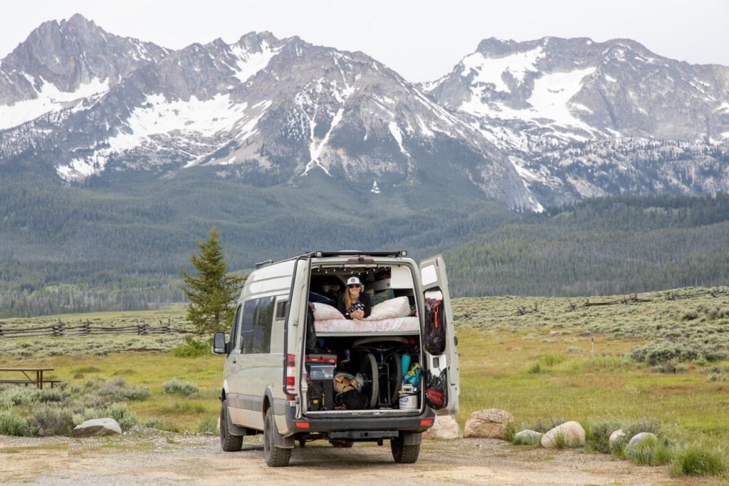 Kristen in her Sprinter van looking out the back in front of distant snowy mountains in Idaho