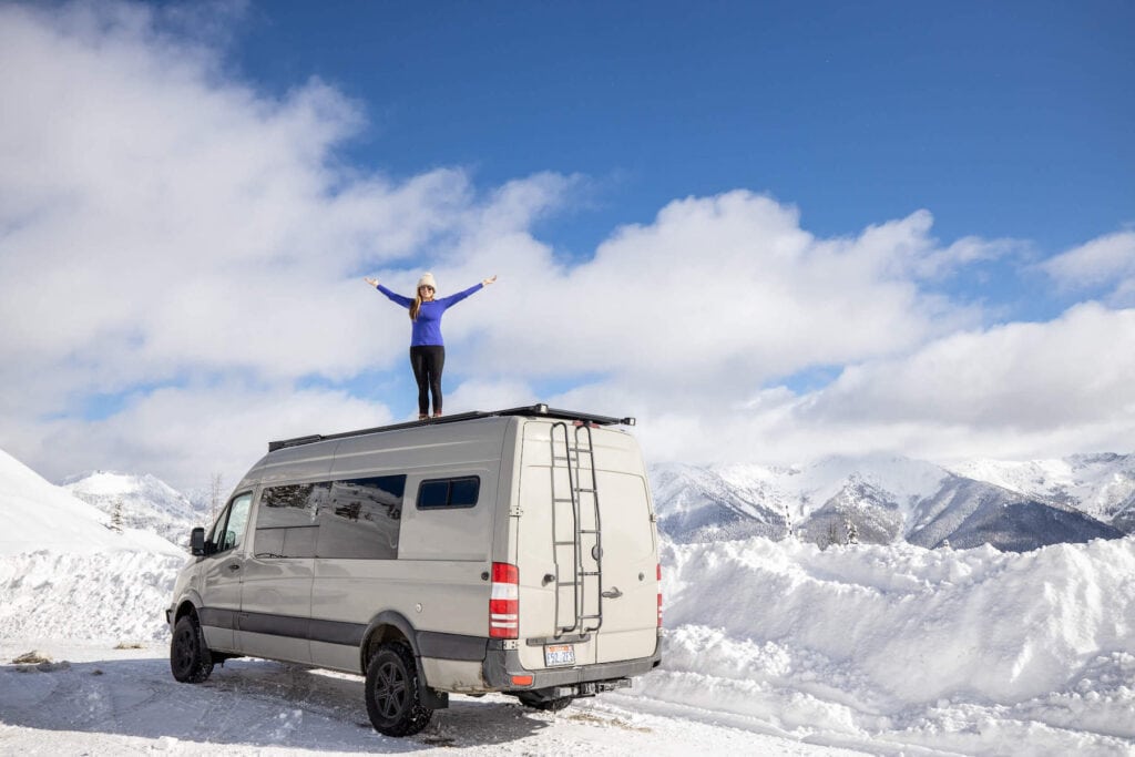 Kristen Bor of the travel blog Bearfoot Theory standing on top of Sprinter van on Galena Summit in Idaho surrounded by snow