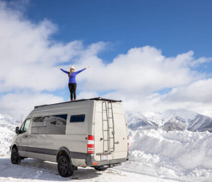 Kristen Bor of the travel blog Bearfoot Theory standing on top of Sprinter van on Galena Summit in Idaho surrounded by snow