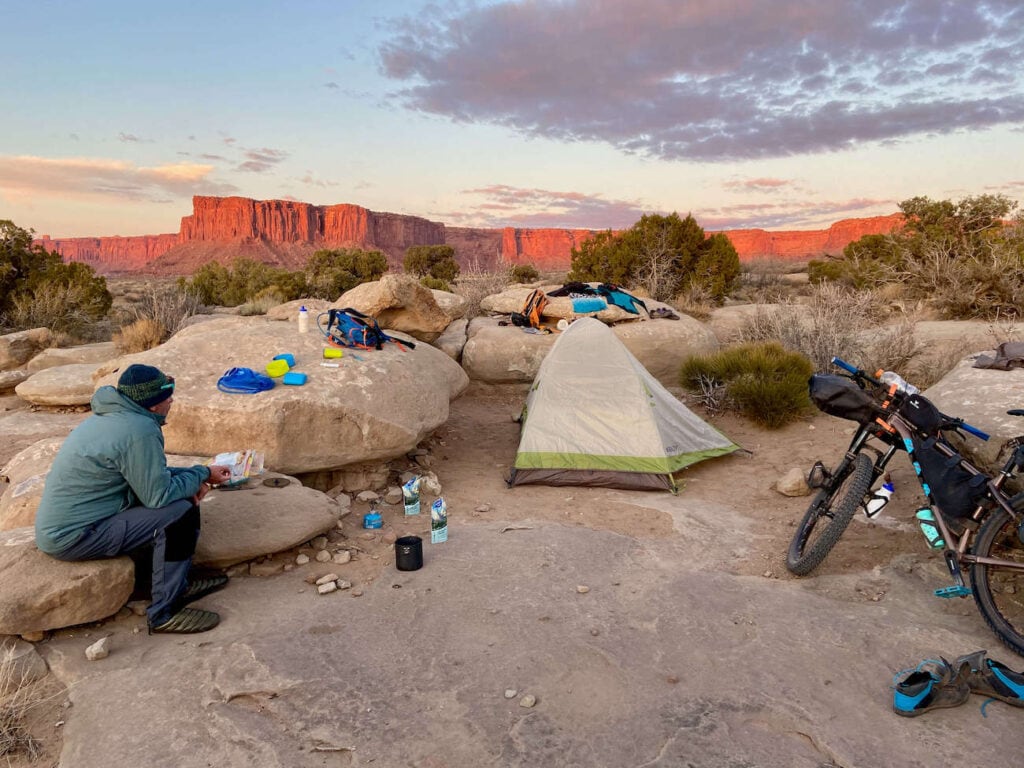 Man sitting on rock at remote desert campsite in Utah with sun setting on red rock bluffs in background. Tent is set up and camping gear is laid out on rocks. Loaded bikepacking bike leaning against boulder. 