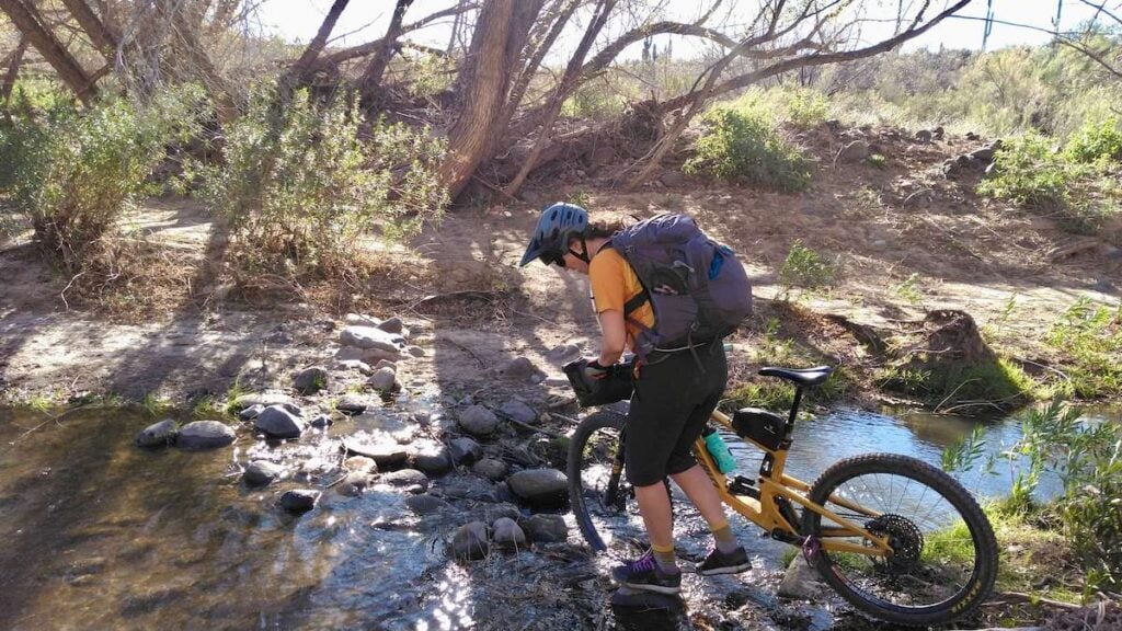 Female mountain biker with large backpack on back pushing bike across narrow and shallow river on overnight bikepacking trip