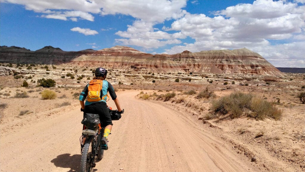 Female rider on fully loaded bikepacking bike pedaling down dirt road in Capitol Reef National Park in Utah with red and white bluffs in the distance