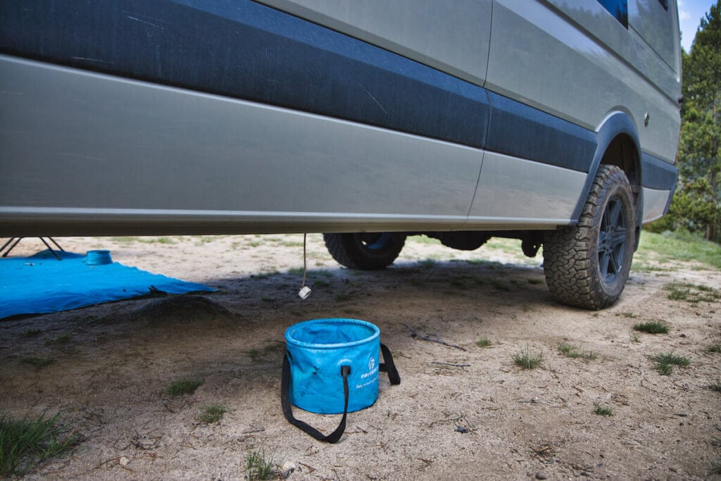 a blue graywater bucket sitting on the ground at a campsite underneath a Sprinter van with a graywater hose sticking out the bottom of the van