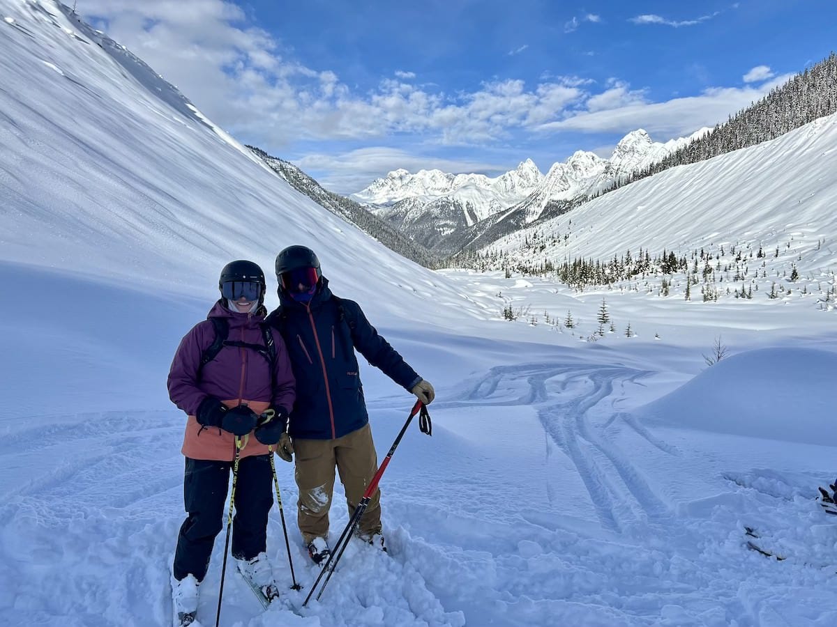 Two people standing for photo on snowy ski slope dressed in skiing gear and equipment