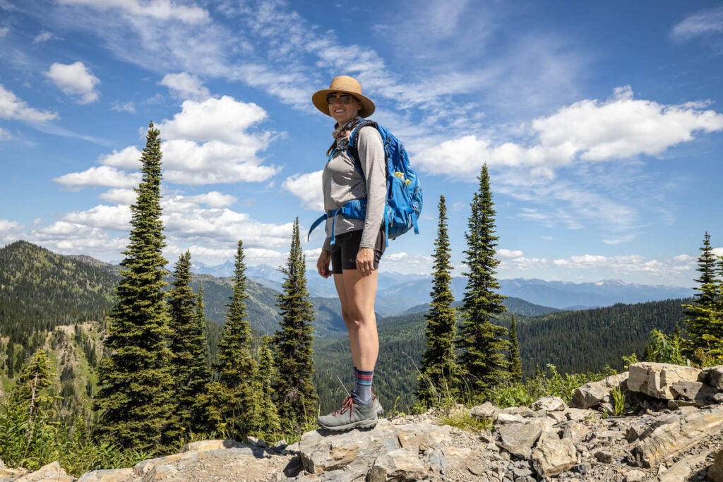 Kristen wearing mid-ankle Oboz Sypes hiking boots on rocky trail in Montana. Pine trees and mountain ranges in the background. 