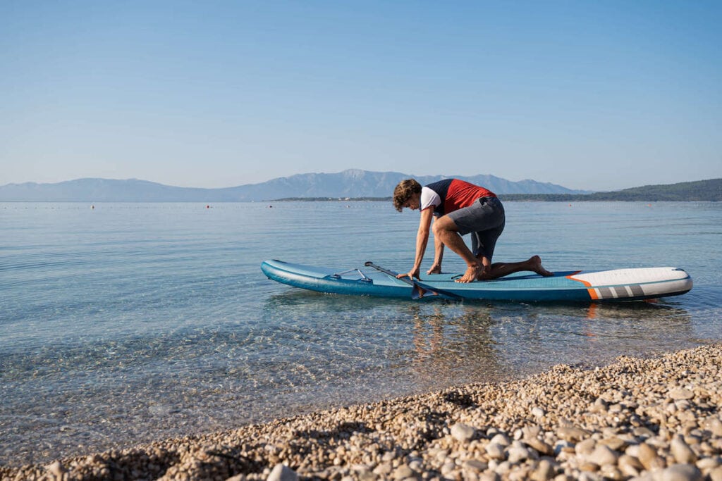 Young man starting to stand up on paddle board in shallow water near shore